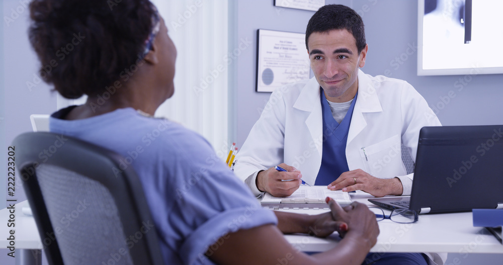 Obraz premium Happy senior patient talking with doctor in medical office about good health. Positive African woman meeting with young latino doctor to discuss her current health condition