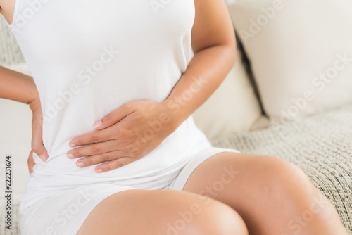 Young woman having painful stomachache and back pain sitting on sofa at home. Chronic gastritis. Abdomen bloating concept.