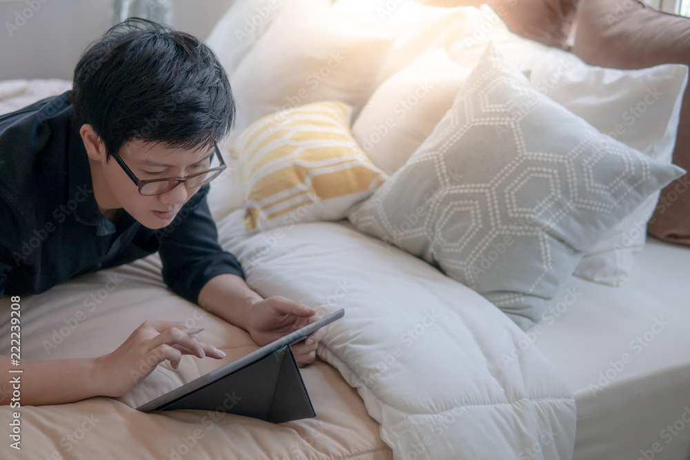 Young Asian business man using digital tablet and pen while lying on bed in cozy bedroom. Home living lifestyle with modern electronic gadget concept Young Asian business man using digital tablet and pen while lying on bed in cozy bedroom. Home living lifestyle with modern electronic gadget concept