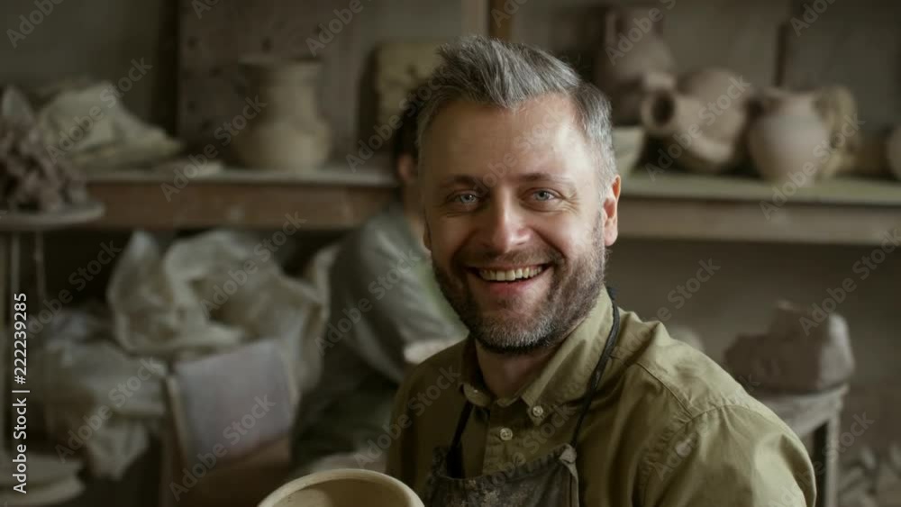 PAN shot of happy bearded man holding ceramic pot and laughing while posing for camera in pottery workshop; cheerful young woman making pottery in wheel in background PAN shot of happy bearded man holding ceramic pot and laughing while posing for camera in pottery workshop; cheerful young woman making pottery in wheel in background