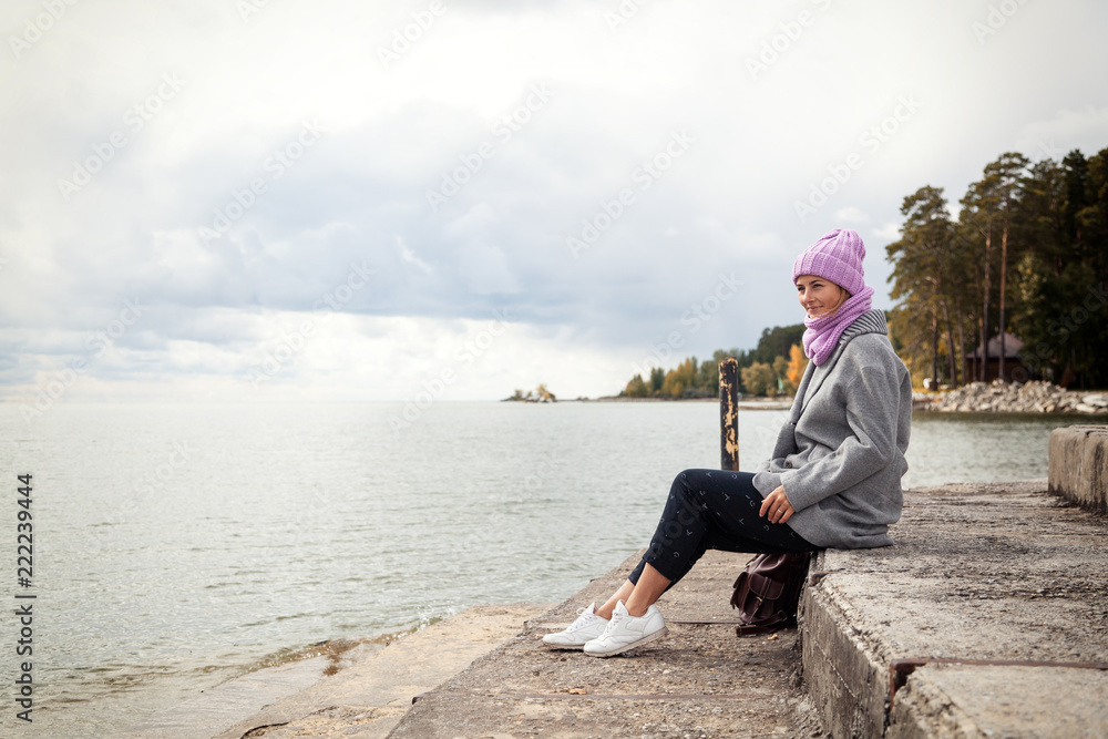 Outdoor atmospheric lifestyle photo of young beautiful  darkhaired woman  in knitting hat, in a gray coat and black trousers sits on the pier next to the sea  in sunny autumn day