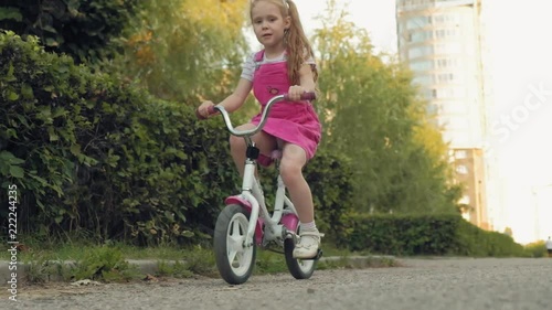 A happy, beautiful, little girl with long blond hair in a pink skirt and jumper rides a children's bike on the road, she smiles. Super slow motion