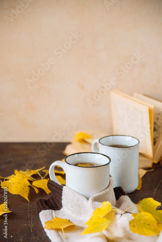two white cups with tea in yellow leaves with a book and a knitted scarf, the concept of warmth and autumn sadness