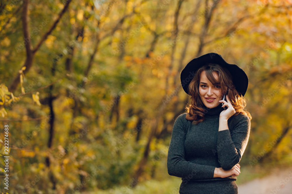 beautiful girl in a black hat holds a phone in her hand in the park in the autumn. Woman walk in the forest along the road beautiful girl in a black hat holds a phone in her hand in the park in the autumn. Woman walk in the forest along the road