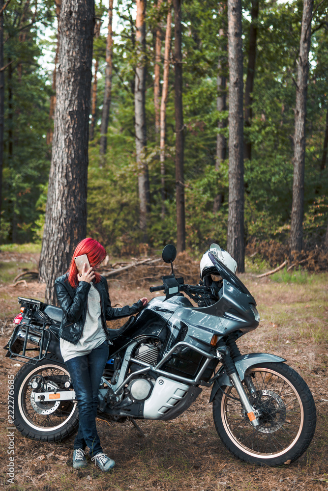 Portrait of happy woman talking on phone, with red hair, dressed in a leather jacket and jeans, stands with adventure tourist motorcycle, in the forest, autumn day, off road travel vertical photo