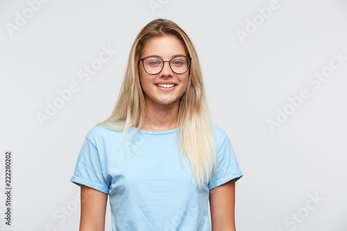 Portrait of happy attractive young woman with blonde hair wears glasses and blue t shirt smiling and looking directly in camera isolated over white background