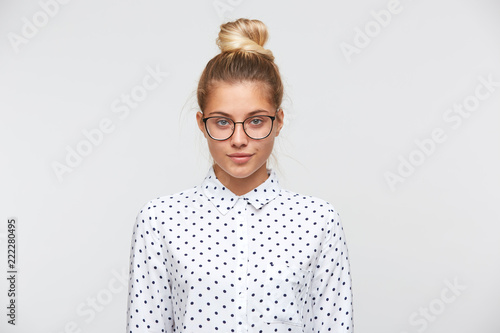 Portrait of confident attractive young woman with bun wears polka dot shirt and spectacles feels serious and looks to the camera isolated over white background