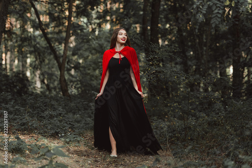 beautiful mystic girl in black dress and red cloak walking in forest