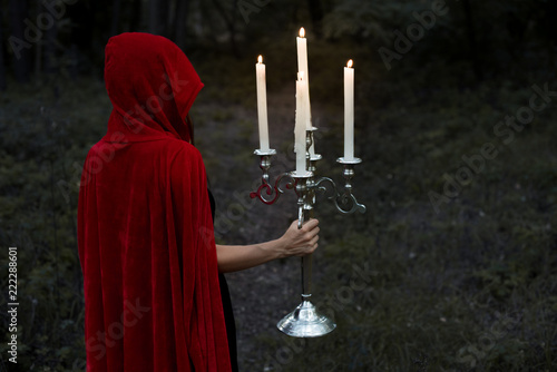 mystic girl in red cloak holding candelabrum with flaming candles in dark woods