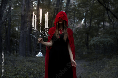 mystic girl in red cloak and hood holding candelabrum with candles in dark forest