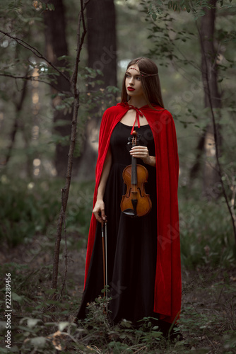 elegant beautiful woman in black dress and red cloak holding violin in forest