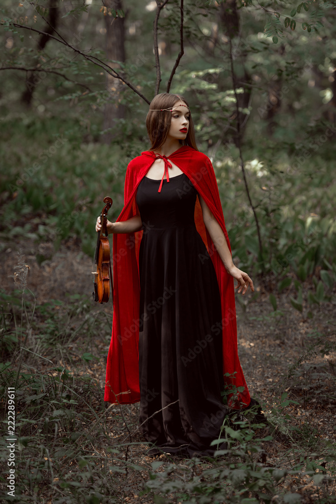 elegant mystic girl in black dress and red cloak holding violin in dark woods
