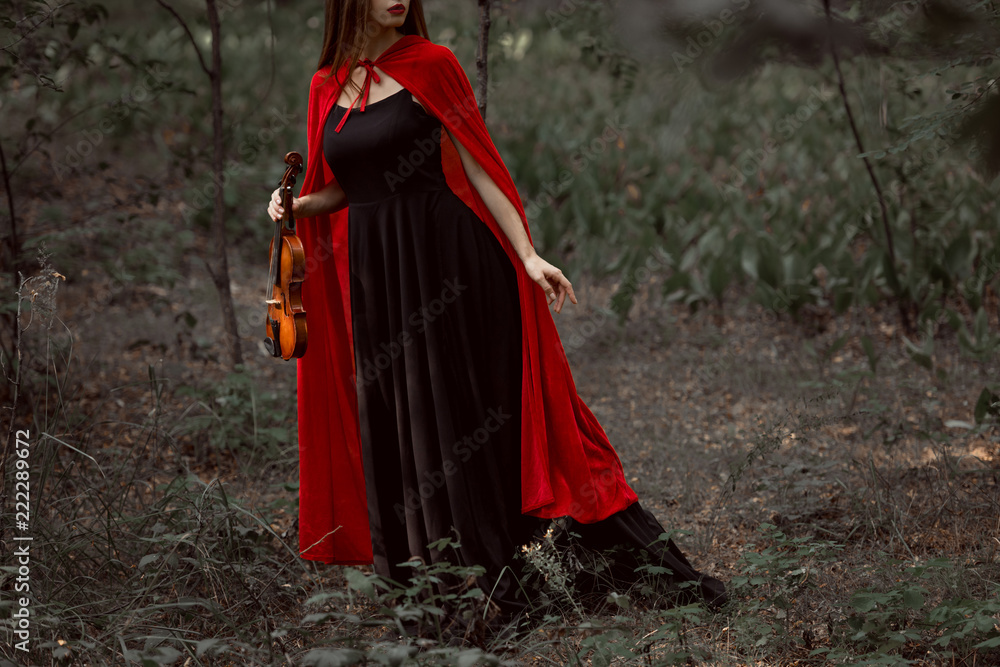 cropped view of elegant mystic woman in black dress and red cloak holding violin in forest cropped view of elegant mystic woman in black dress and red cloak holding violin in forest