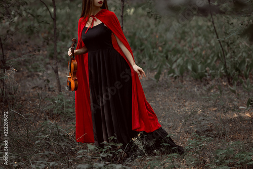 cropped view of elegant mystic woman in black dress and red cloak holding violin in forest