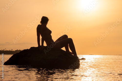 Silhouette of a beautiful woman sitting on a rock at sunset looking at the pacific ocean in summer in Puerto Rico © dennisvdwater