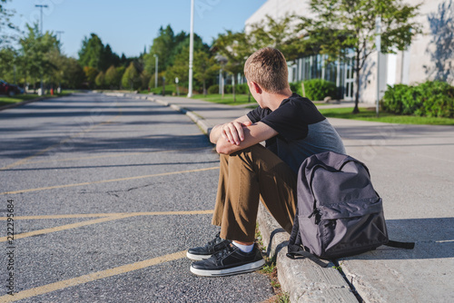 Lonely teenage boy sitting on curb next to high school. 