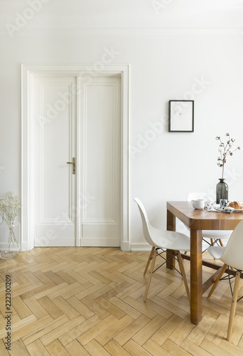 A bright dining room interior with a wooden table and chairs standing on a herringbone parquet against a white wall with double door. Real photo. photo