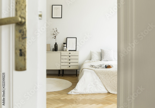 A peek through a half open door into a monochromatic beige and white bedroom interior with a bed and a drawer cabinet. Real photo. photo