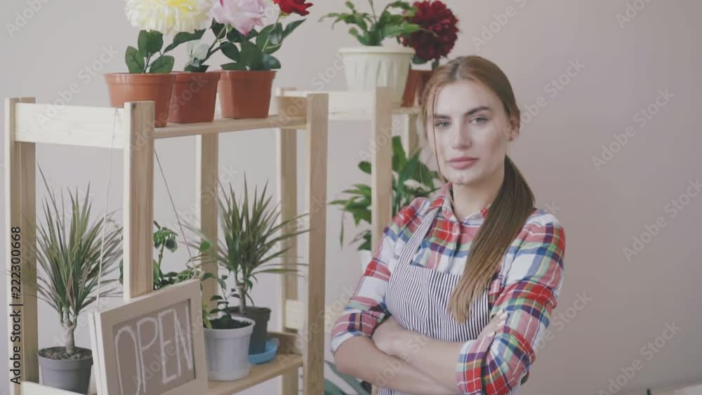 Small business. A girl in a striped apron and plaid shirt stands in a flower shop. On the shelves are flowers and an open sign. Girl florist standing near the shelf with flowers. Small business. A girl in a striped apron and plaid shirt stands in a flower shop. On the shelves are flowers and an open sign. Girl florist standing near the shelf with flowers.