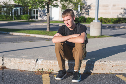 Sad teenage boy sitting on the curb in front of his school.