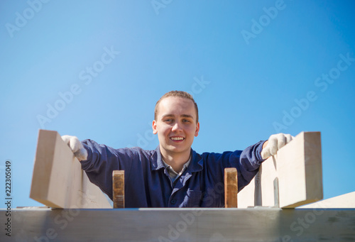 A smiling young man in gloves and work clothes is building a roof