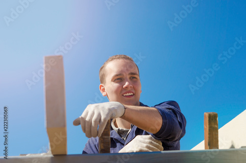 A smiling young man in gloves and work clothes is building a roof