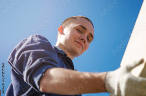 A smiling young man in gloves and work clothes is building a roof