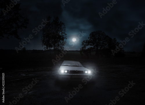 A deserted white car with lights on in a mysterious wilderness. Night scene with full moon.
