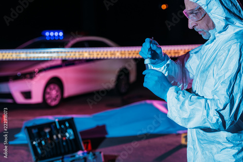 focused male criminologist in protective suit and latex gloves putting evidence into flask by tweezers at crime scene with corpse photo