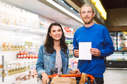Young smiling couple with shopping list and trolley happily looking in camera in dairy department of supermarket