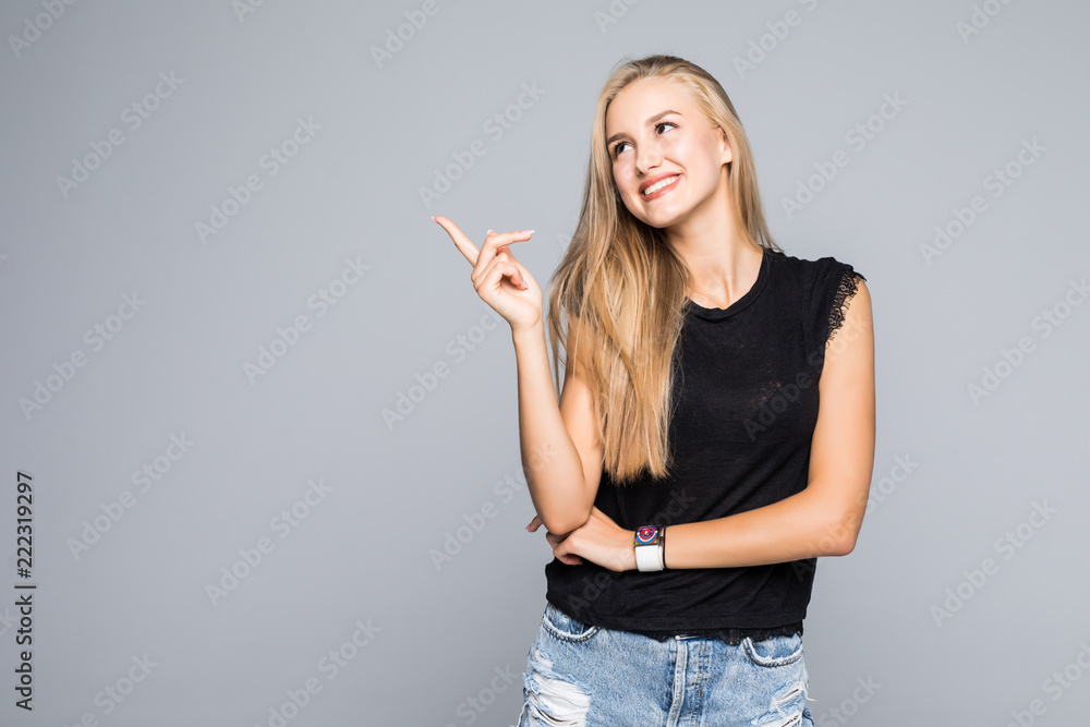 Portrait of a cheerful and happy young beautiful woman in a black T-shirt looking with a smile at the camera and pointing a finger to the side isolated on the gray background. Portrait of a cheerful and happy young beautiful woman in a black T-shirt looking with a smile at the camera and pointing a finger to the side isolated on the gray background.