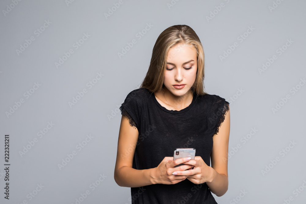 Surfing the net. Attractive young woman smiling and using her smart phone while standing against grey background Surfing the net. Attractive young woman smiling and using her smart phone while standing against grey background