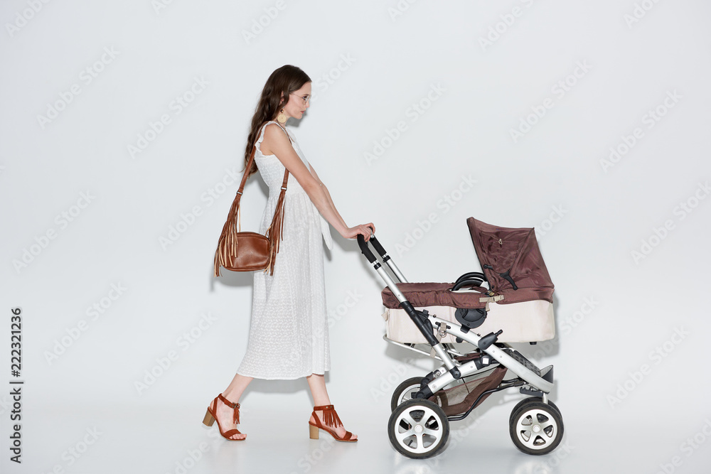 side view of woman in stylish white dress walking with baby stroller on grey side view of woman in stylish white dress walking with baby stroller on grey