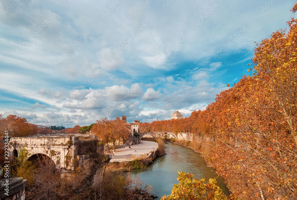 Autumn and foliage in Rome. Red and yellow leaves near Tiber Island with two ancient bridges and beautiful sky, in the city historic center