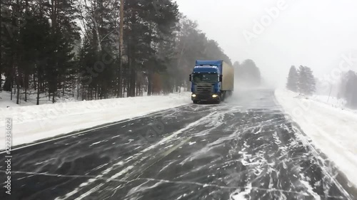  The car is on a snow-covered highway. Heavy truck with trailer carries cargo.  Winter road with a gusty wind and a snowstorm, slow motion. 