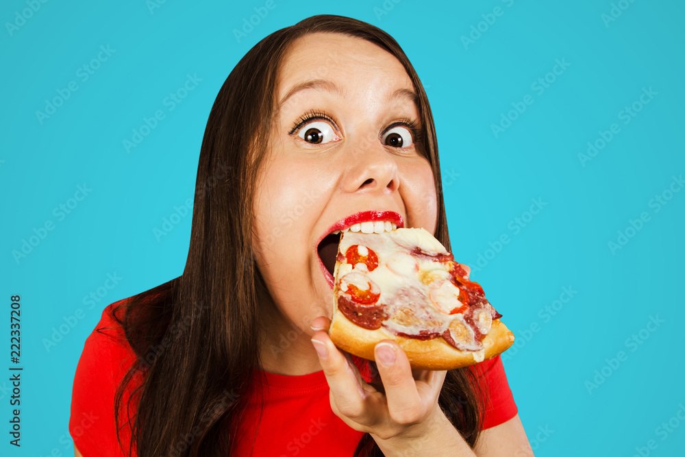 Young pretty girl eats piece of pizza with tomatoes, salami and cheese isolated on a blue background. Young pretty girl eats piece of pizza with tomatoes, salami and cheese isolated on a blue background.