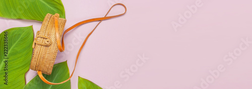 Fashionable handmade natural round rattan bag and tropical leaves on pink background flat lay. Top view with copy space. Trendy bamboo bag Ecobags from Bali. Summer fashion concept Long banner