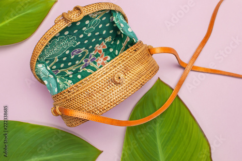 Fashionable handmade natural round rattan bag and tropical leaves on pink background flat lay. Top view with copy space. Trendy bamboo bag Ecobags from Bali. Summer fashion concept. photo