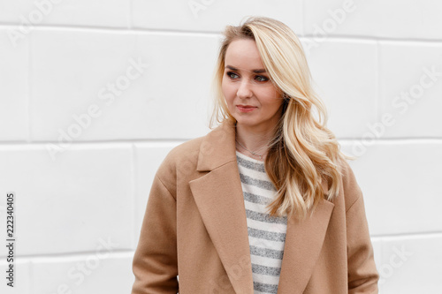 Beautiful young stylish blonde woman wearing beige coat and striped jumper posing near white street wall. Trendy casual outfit. Street fashion.