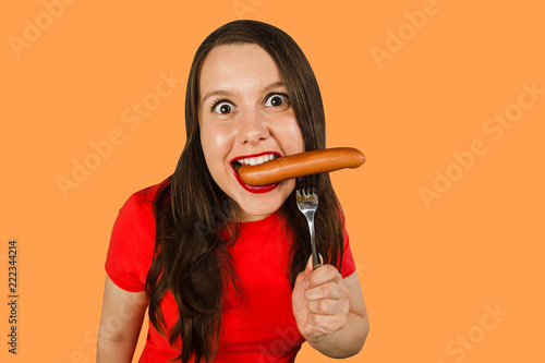 Young cute girl eating sausage on a fork at her mouth, isolated on orange background