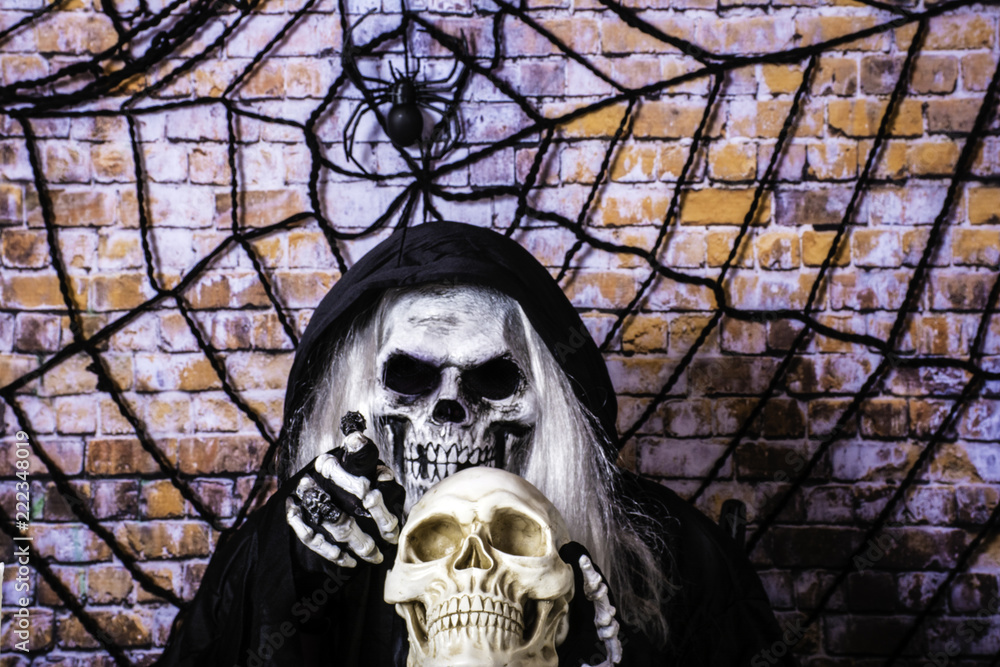 man in black Grimm reaper robe and skull mask pointing at camera with  skeleton hands holding a human skull with brick wall background covered by  large spider and web Stock Photo