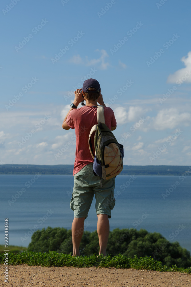 A traveler with a backpack behind him is standing on a hill and taking a picture of the lake.