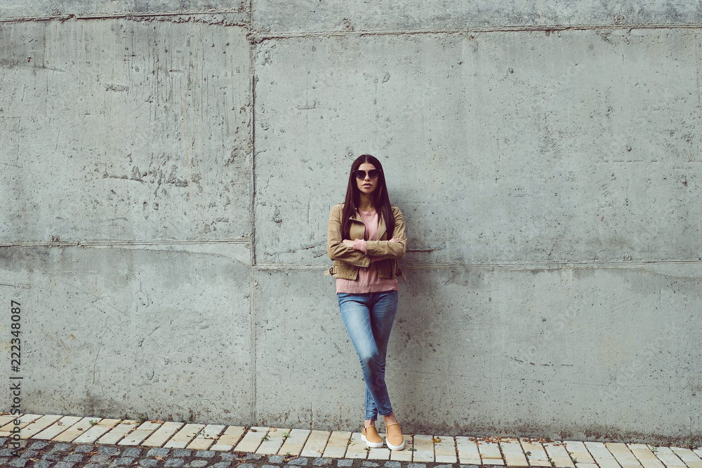 Casual and calm. Full length of beautiful young woman looking at camera while standing against concrete background outdoors Casual and calm. Full length of beautiful young woman looking at camera while standing against concrete background outdoors