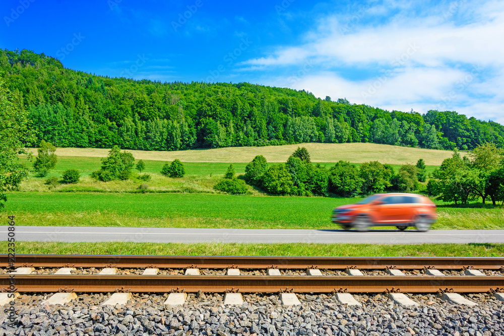 A fast moving car along the road goes alongside the railway tracks. Tracks running next to the road. A sunny view of a car passing through the countryside