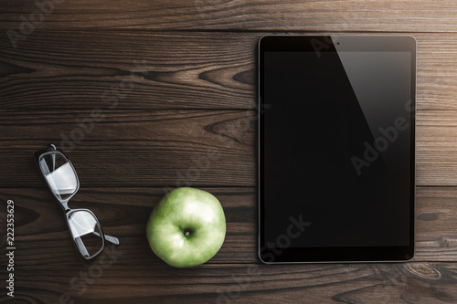 Tablet black screen for advertisement display, apple and eyeglasses on wood background. Top view, light effect