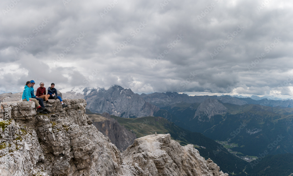 four young male and female hikers sitting on a mountain peak ledge in the Dolomites and looking at the amazing view with Langkofel and Marmolada peaks