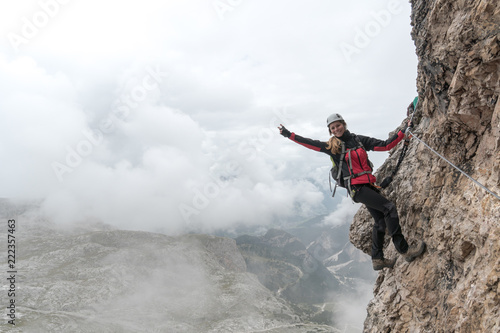 young attractive female university student on a vertical and exposed rock face climbs a Via Ferrata while smiling and waving and making a peace sign
