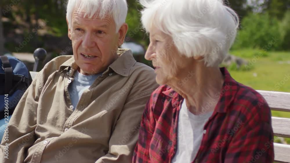 Senior man and woman sitting on bench in national park and talking while taking a break during hike at summer day Senior man and woman sitting on bench in national park and talking while taking a break during hike at summer day