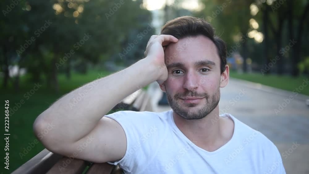 Attractive young man sitting on a park bench with beautiful smile on his face, he is on a date enjoying a conversation. Sexy guy in white t shirt with a hot look smiling. he knows how to seduce woman Attractive young man sitting on a park bench with beautiful smile on his face, he is on a date enjoying a conversation. Sexy guy in white t shirt with a hot look smiling. he knows how to seduce woman