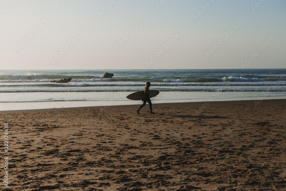 young surfer man walking by the sea shore holding his surf table. Sunset. Summertime, sport and holidays concept